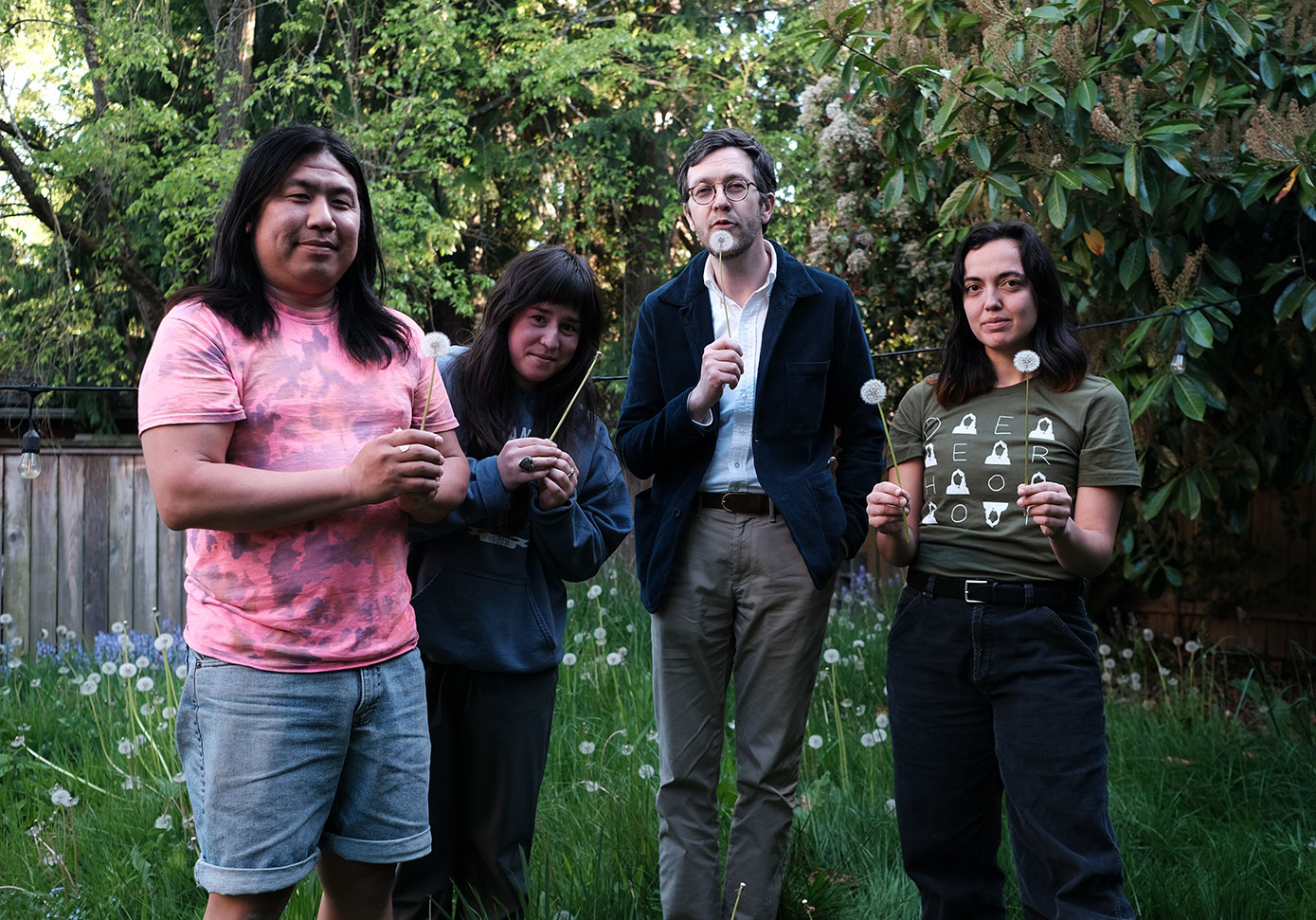 Evan, Izumi, Craig, and Kirsten holding dandelions in a lush dandelion-filled yard.  Craig is very cutely blowing his dandelion.